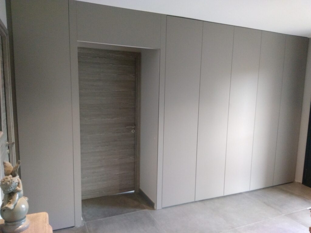 Agencement sur mesure consistant à couvrir intégralement le mur de rangements, fermés du sol au plafond par des portes à système touche-lâche autour d'une porte